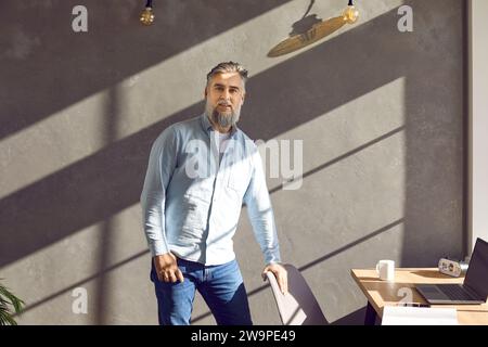 Reifer Geschäftsmann, der in der Nähe seines Arbeitsplatzes auf einem grauen Hintergrund steht. Stockfoto