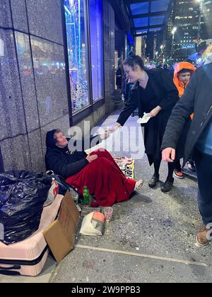 Im Weihnachtsgeist gibt eine Frau ein paar Dollar an eine obdachlose ältere Frau, die während der Weihnachtsfeiertage in New York City auf dem Bürgersteig entlang der 5th Avenue sitzt. Stockfoto