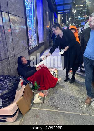 Im Weihnachtsgeist gibt eine Frau ein paar Dollar an eine obdachlose ältere Frau, die während der Weihnachtsfeiertage in New York City auf dem Bürgersteig entlang der 5th Avenue sitzt. Stockfoto