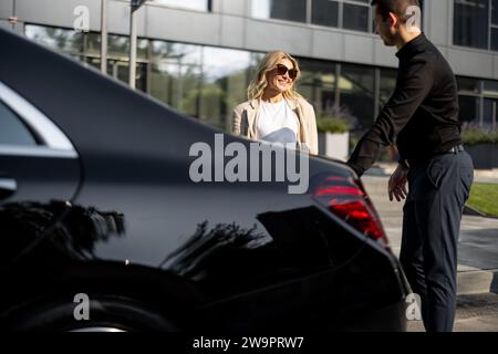 Geschäftsfrau wartet, während der Chauffeur einen Koffer in den Kofferraum packt und während einer Geschäftsreise ein luxuriöses Taxi nutzt. Konzept des Business Transfer Service Stockfoto