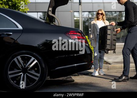 Chauffeur packt einen Koffer in einen Kofferraum, Geschäftsfrau wartet in der Nähe mit einem luxuriösen Taxi während einer Geschäftsreise. Konzept des Unternehmensübertragungskonzepts Stockfoto