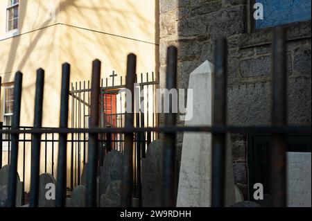Salem, Massachusetts – Ein kleines hinteres Eisenkreuz auf der Spitze eines Friedhofstors wird in spätem Sonnenlicht gewaschen, während der kleine Friedhof im Vorfeld liegt Stockfoto