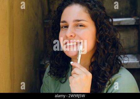 Nahaufnahme einer jungen Latina-Frau mit Lockenwicklern, nachdenklich lächelnd und in die Kamera schauend, den Stift mit der Hand über ihr Gesicht hielt. Stockfoto