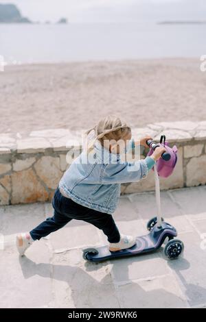 Das kleine Mädchen reitet mit einem Roller auf einem gekachelten Pfad entlang der Küste und stößt mit dem Fuß davon Stockfoto