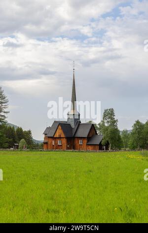 Eine vertikale Aufnahme der alten Kvikne kirke mit ihrem hohen Turm, eingebettet in Grün vor einem wolkenübersäten Himmel in Kvikne, Innlandet Stockfoto