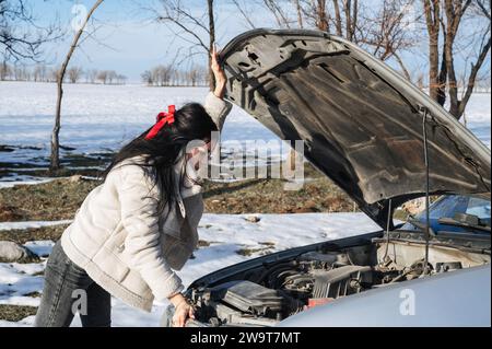 Mädchen schaut unter der Motorhaube auf den Motor eines kaputten Autos auf der Straße im Winter Stockfoto