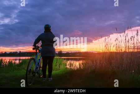 Mädchen mit Rennrad steht am Ufer eines Sees vor einem wunderschönen Himmel bei Sonnenuntergang. Das Konzept eines aktiven Lebensstils. Stockfoto