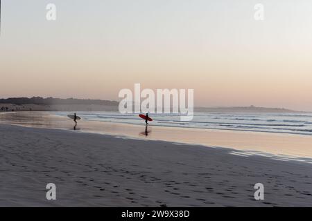 Zwei Surfer laufen bei Sonnenuntergang mit Windturbinen am Ufer entlang in Essaouira, Marokko. 30. Dezember 2023 Stockfoto