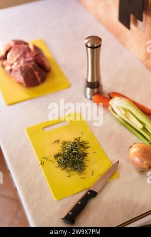 Fein gehackter Rosmarin auf einem Schneidebrett liegt neben rohen Filet und Gemüse auf dem Tisch Stockfoto