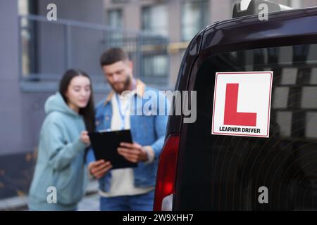 Fahrer und Kursleiter mit Klemmbrett in der Nähe des Autos im Freien, selektiver Fokus auf L-Platte. Fahrschule Stockfoto