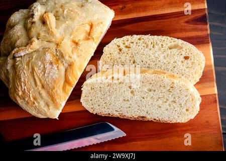 Scheiben frisch zubereitetes, knuspriges französisches Brot: Geschnittenes Stück hausgemachtes französisches Brot auf einem hölzernen Schneidebrett mit einem gezackten Brotmesser Stockfoto