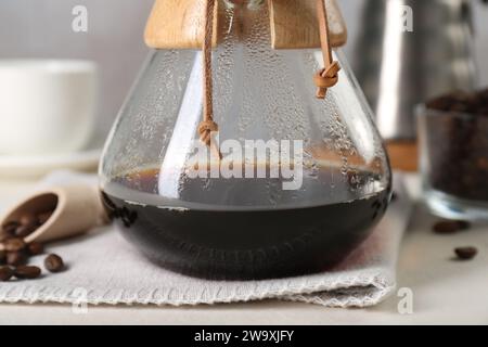 chemex-Glaskaffeemaschine mit leckerem Tropfkaffee und Bohnen auf weißem Tisch, Nahaufnahme Stockfoto