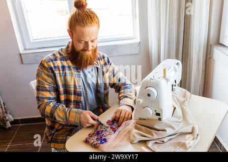 Hübscher rothaariger Mann mit langen Bartnähten an einer Nähmaschine im Studio Stockfoto