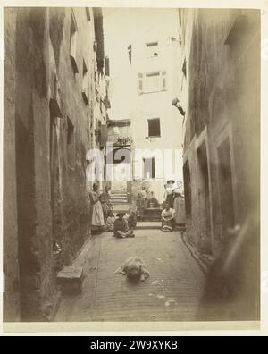 Eine enge Straße in Menton mit Frauen und Kindern in den Straßen, ca. 1886 - ca. 1896 Fotografieren Sie Eine enge Arbeiterstraße in Menton mit Kindern und Frauen in Arbeitskleidung. Hinter der Straße befindet sich ein Wasserpunkt, an dem die Menschen Eimer mit Wasser füllen. Unterstützung für die Fotografie in Frankreich. Pappe mit Albumendruck Kin Stockfoto