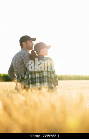 Ein paar Bauern in karierten Hemden und Mützen stehen bei Sonnenuntergang auf einem landwirtschaftlichen Weizenfeld. Stockfoto
