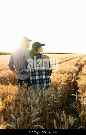 Ein paar Bauern in karierten Hemden und Mützen stehen bei Sonnenuntergang auf einem landwirtschaftlichen Weizenfeld. Stockfoto