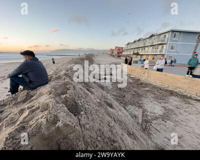 30. Dezember 2023, Santa Barbara/Ventura, Kalifornien, USA: 30. Dezember: Ein Mann fuhr eine Stunde, um bei Sonnenuntergang bei Ebbe nach Pierpont, Ventura zu kommen, um zu sehen, wo ein â œrouge waveâ€ den Ozean durch die Straßen gewaschen hatte. Hinter ihm sind Dutzende von anderen Menschen aus ganz Südkalifornien, die hierher kamen, um dem Tatort der oceanâ zu huldigen. Die Sanddüne wurde von den Crews des County of Ventura gebaut, um zu verhindern, dass weitere Häuser und Geschäfte beschädigt werden: Es werden mehr große Wellen und Überschwemmungen erwartet, besonders bei Flut. Die Strassen zum Strand sind alle geschlossen Stockfoto