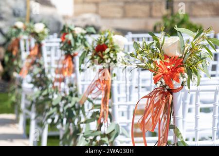 Weiße Holzstühle, dekoriert mit Blumen und üppigem Grün als Teil einer Hochzeitszeremonie, akribische Vorbereitung des Außenbereichs Stockfoto