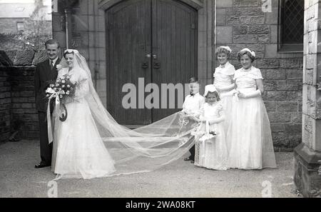 1950er Jahre, historisch, Hochzeit, außerhalb der Kirche, halten ein kleines Pökelchen und ein junges Blumenmädchen den Zug der Braut, während sie mit dem neuen Ehemann, England, Großbritannien, steht, mit ihren zwei Brautjungfern. Stockfoto