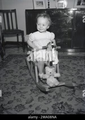 1960er Jahre, historisch, in einem vorderen Roon, ein kleines Mädchen, das auf einem hölzernen Schaukelpferd reitet, England, Großbritannien, ihr Teddybär auf der Vorderseite, der eine Fahrt macht! Stockfoto