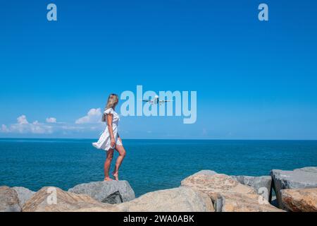 Küstenmagie: Mädchen in weißem Kleid auf Steinen, treffen ein Flugzeug am Blauen Meer Stockfoto