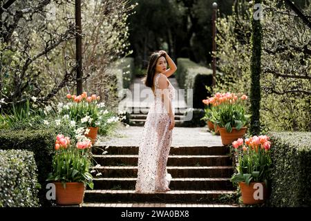 Schöne Passform kurvige junge asiatische Frau transparentes Paillettenkleid stehend in einem Garten Tulpen Pink Stockfoto