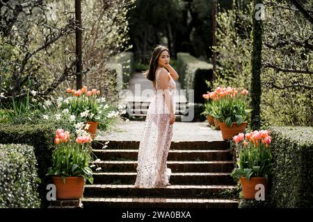 Schöne Passform kurvige junge asiatische Frau transparentes Paillettenkleid stehend in einem Garten Tulpen Pink Stockfoto