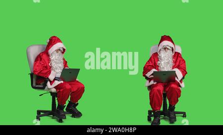 Der moderne saint Nick arbeitet auf einem Laptop im Studio und sitzt auf einem Stuhl vor dem Ganzkörper-Grünschirm-Hintergrund. weihnachtsmann-Figur mit schnurlosem, tragbarem pc, Online-Website im festlichen Kostüm durchsuchen. Stockfoto