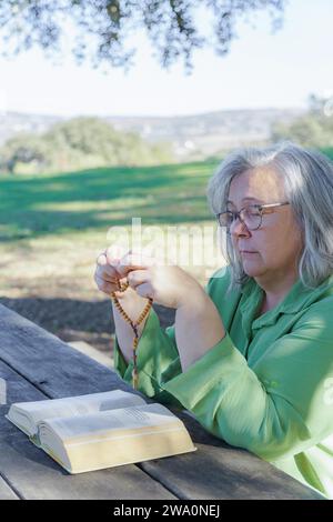 Reife weißhaarige Frau mit Brille betet mit Rosenkranzperlen und einer Bibel auf einem Holztisch auf dem Feld Stockfoto