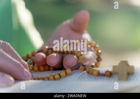 Die Hände einer Frau beten den Rosenkranz auf dem Feld über einer offenen Bibel auf einem Holztisch Stockfoto