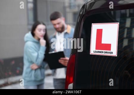Fahrer und Kursleiter mit Klemmbrett in der Nähe des Autos im Freien, selektiver Fokus auf L-Platte. Fahrschule Stockfoto