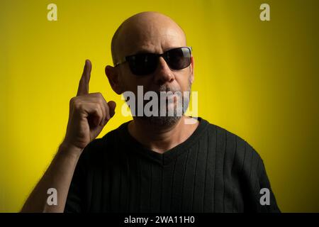 Porträt eines glatzköpfigen, bärtigen, reifen Mannes, der Sonnenbrille trägt und Zeichen mit den Händen macht. Isoliert auf gelbem Hintergrund. Stockfoto