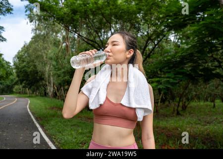 Eine junge asiatische Frau in Fitnesskleidung hält an, um Wasser aus ihrer Plastikflasche zu trinken, während sie morgens auf einer Laufbahn eines Einheimischen trainieren kann Stockfoto