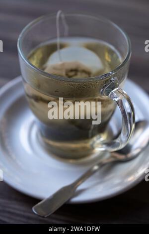 Nahaufnahme eines Teeglases gefüllt mit heißem Wasser und einem Teebeutel darin auf einer weißen Untertasse mit einem Löffel daneben auf einem Holztisch. Geringe Schärfentiefe, Stockfoto