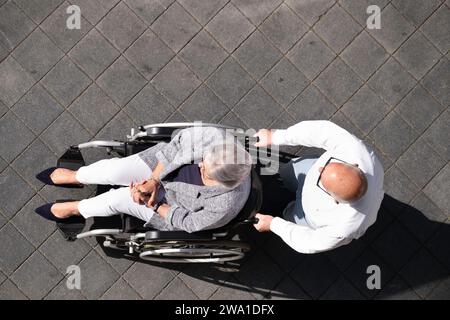 Älteres Paar im Sommer von oben gesehen. Der Mann schiebt seine Frau im Rollstuhl entlang des Bürgersteigs. Kopierbereich Stockfoto