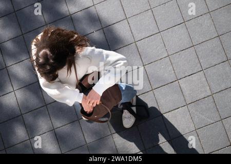 Unerkennbare junge Frau mit weißem Pullover steht auf einem Bürgersteig und schaut in ihre braune Schultertasche. Von oben gesehen, langer Schatten Stockfoto