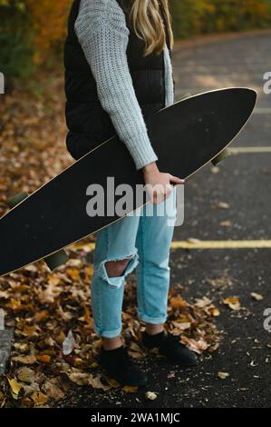 Zugeschnittene Ansicht einer Frau, die Longboard auf einem Parkplatz mit Blättern hält Stockfoto