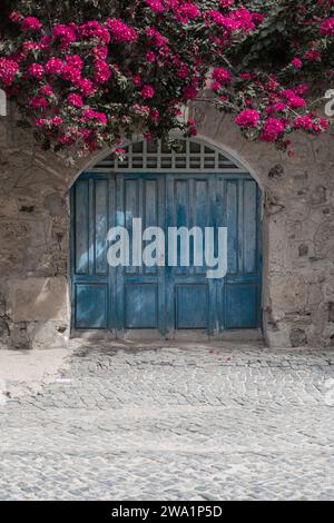 Eine wunderschöne verwitterte blaue Tür, umgeben von sandigen Steinmauern. Mit einer wunderschönen rosafarbenen Blume, die ihn schön umrahmt. Stockfoto