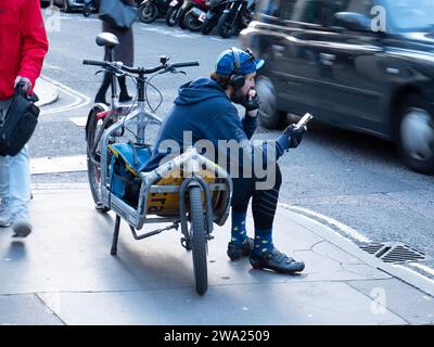 London, Großbritannien. November 2023. Ein Fahrradkurier sitzt auf einem elektrischen Lastenfahrrad und überprüft sein Handy auf einer Straße in Soho nach neuen Lieferaufgaben. Das Bild zeigt die „Gig Economy“-Realität moderner Stadtlogistik, in der Fahrer von Unternehmen wie Pedal Me, Zhero oder CitySprint zwischen den Jobs auf App-basierte Benachrichtigungen warten. Ende 2023 kam es in London zu einem deutlichen Anstieg des Einsatzes von E-Cargo-Fahrrädern, da Unternehmen zunehmend Diesel-Transporter gegen emissionsfreie Alternativen tauschten, um die Ultra Low Emission Zone (ULEZ) und die Staugebühr der Hauptstadt zu befahren Stockfoto