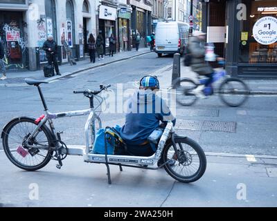 London, Großbritannien. November 2023. Ein Fahrradkurier sitzt auf einem elektrischen Lastenfahrrad und überprüft sein Handy auf einer Straße in Soho nach neuen Lieferaufgaben. Das Bild zeigt die „Gig Economy“-Realität moderner Stadtlogistik, in der Fahrer von Unternehmen wie Pedal Me, Zhero oder CitySprint zwischen den Jobs auf App-basierte Benachrichtigungen warten. Ende 2023 kam es in London zu einem deutlichen Anstieg des Einsatzes von E-Cargo-Fahrrädern, da Unternehmen zunehmend Diesel-Transporter gegen emissionsfreie Alternativen tauschten, um die Ultra Low Emission Zone (ULEZ) und die Staugebühr der Hauptstadt zu befahren Stockfoto