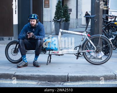 London, Großbritannien. November 2023. Ein Fahrradkurier sitzt auf einem elektrischen Lastenfahrrad und überprüft sein Handy auf einer Straße in Soho nach neuen Lieferaufgaben. Das Bild zeigt die „Gig Economy“-Realität moderner Stadtlogistik, in der Fahrer von Unternehmen wie Pedal Me, Zhero oder CitySprint zwischen den Jobs auf App-basierte Benachrichtigungen warten. Ende 2023 kam es in London zu einem deutlichen Anstieg des Einsatzes von E-Cargo-Fahrrädern, da Unternehmen zunehmend Diesel-Transporter gegen emissionsfreie Alternativen tauschten, um die Ultra Low Emission Zone (ULEZ) und die Staugebühr der Hauptstadt zu befahren Stockfoto