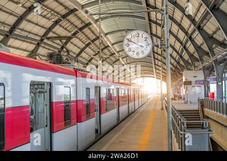 Uhr an einem Pfosten auf dem Fahrgastbahnsteig eines Bahnhofs, die auf die Abfahrt eines elektrischen Pendlerzuges wartet, der auf Passagiere mit geöffneten Türen wartet Stockfoto