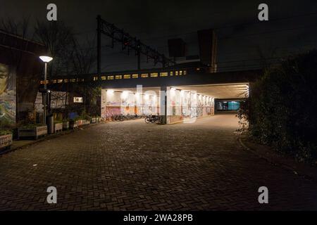 Nachtblick auf beleuchtete Tunnel (Unterführungen) unter Bahngleisen, auf denen der Zug vorbeifährt, mit geparkten Fahrrädern und Graffiti an den Wänden Stockfoto