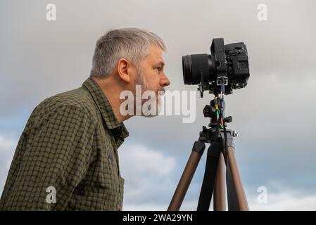 Fotograf, graues Haar und Vollbart, 56 Jahre alt, blickt in seine 35-mm-Spiegelreflexkamera auf dem Stativ, grau bedeckter Himmel Stockfoto