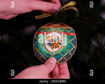 Eine Frau, die ein kreuzgenähtes zeitloses Weihnachtsschmuck an einen Weihnachtsbaum hängt. Dieses grüne Weihnachtsschmuck mit Horn, Pflanze und Bogen e Stockfoto