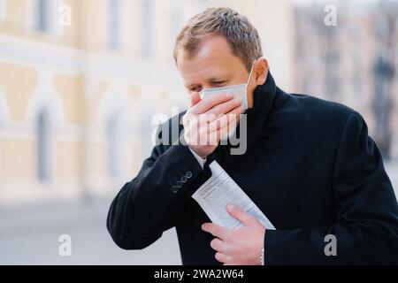 Kranker Mann in schwarzem Fell, der den Mund mit Maske bedeckt, draußen, Anzeichen von Krankheit, in einer städtischen Umgebung, die gesundheitliche Bedenken hervorhebt Stockfoto