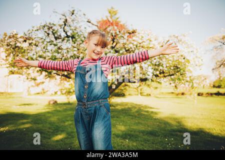 Frühlingsporträt eines süßen kleinen Mädchens, das an einem schönen sonnigen Tag im blühenden Garten spielt, mit Denim-Overall und Streifenhemd Stockfoto