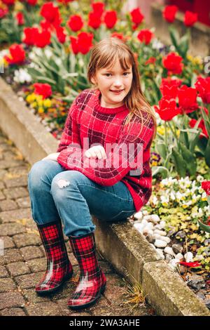 Frühlingsportrait im Freien: Ein entzückendes Mädchen mit rotem kariertem Pullover und Regenstiefeln, ein Kind, das an einem regnerischen Tag draußen spielt, Mode für Mädchen Stockfoto