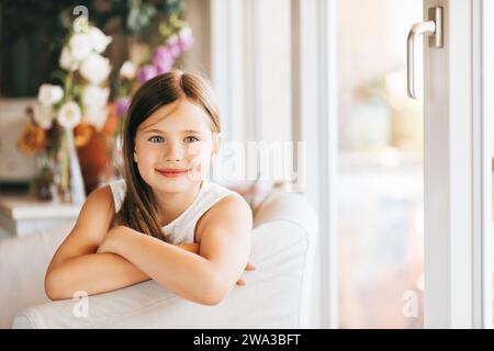 Niedliches kleines Mädchen, das zu Hause in einem weißen Stuhl neben dem Fenster sitzt Stockfoto