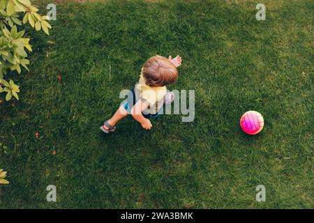 Kleiner Junge, der draußen mit dem Ball auf grünem Rasen spielt, Blick von oben Stockfoto
